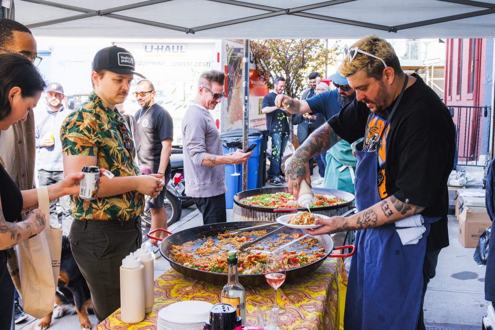 Chef plating from large paella pans under a tent while guests wait on the sidewalk