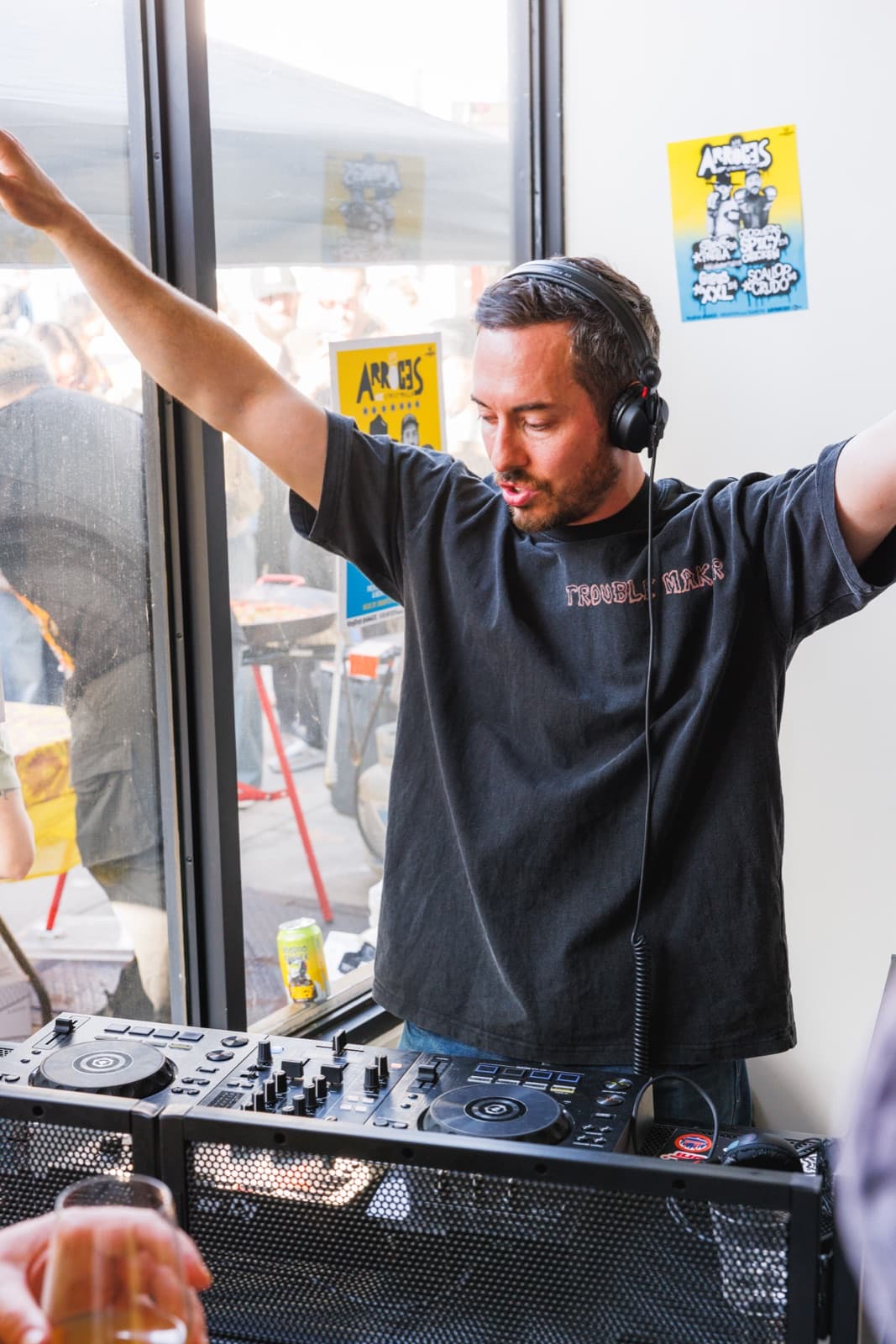 DJ performing indoors with a paella pan and crowd visible through the window behind him