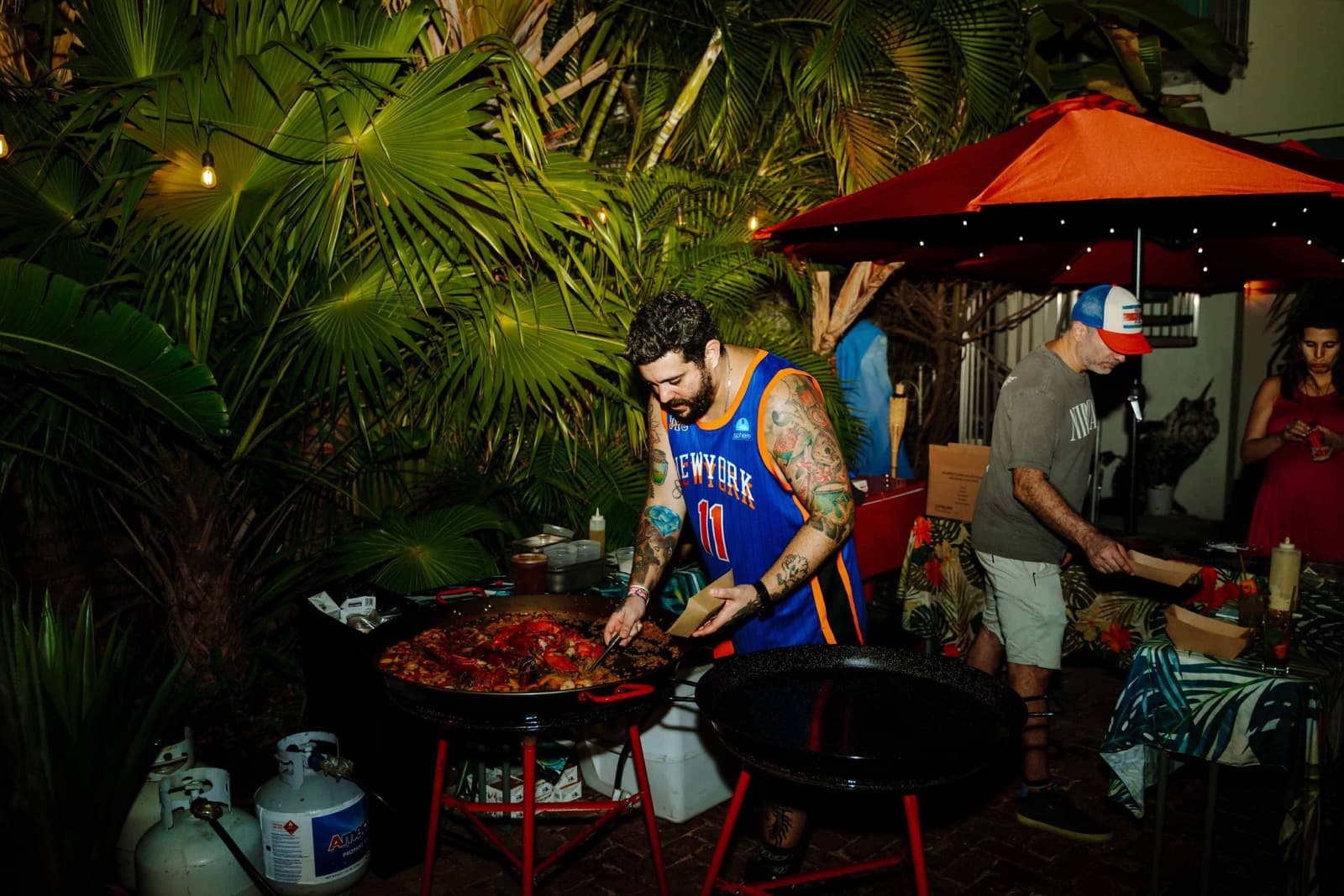 Chef Eduardo in a Knicks jersey serving lobster paella from a large pan at an outdoor night pop-up