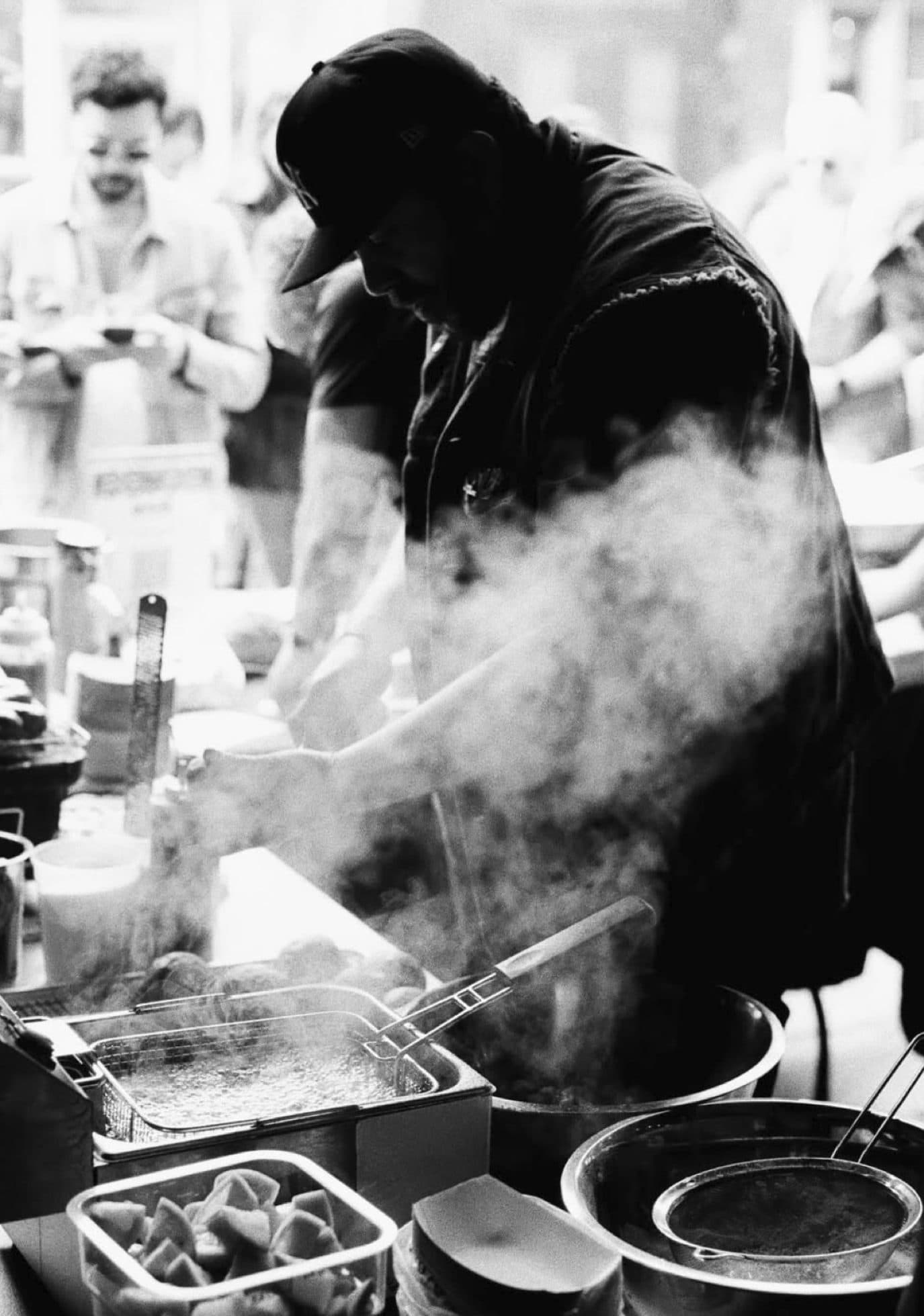 Chef Eduardo Cuenca in profile cooking at a busy outdoor pop-up, steam rising from the station